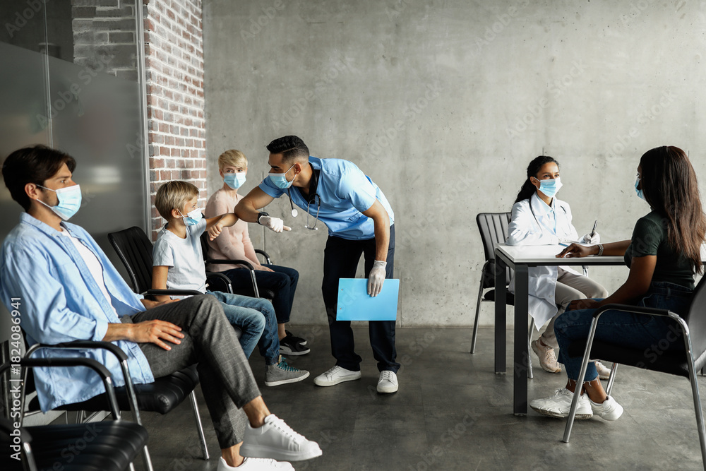 © Prostock-studio - Middle eastern young man doctor greeting teen boy patient with elbow bump, multiracial group of people sitting at clinic, waiting for vaccination against COVID-19, wearing protective face masks