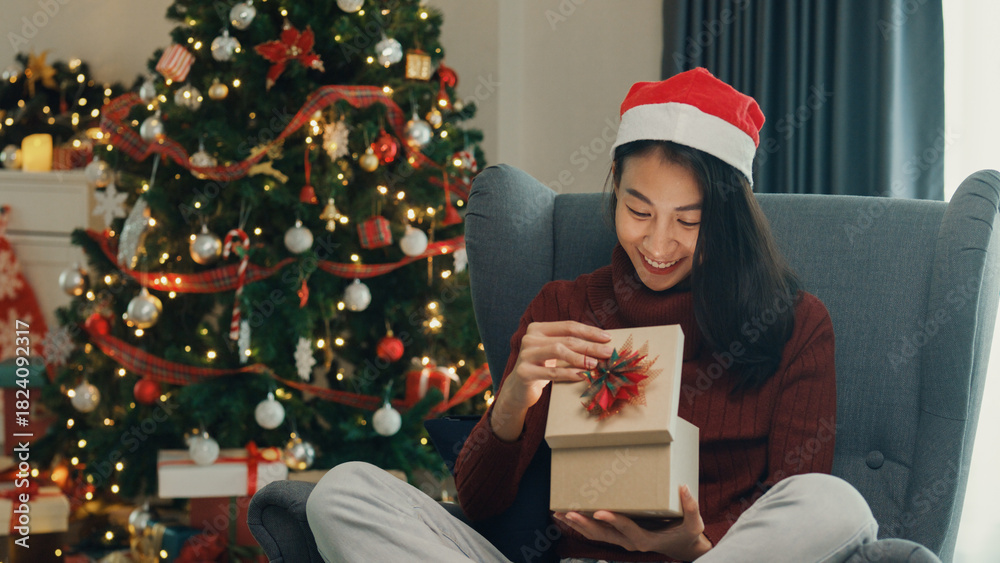 Fototapeta premium Surprise excited Asia female wear red sweater new normal celebrate christmas eve unboxing online shopping present gift delivery from friend and family on wing chair in living room at house.