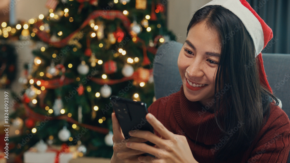 Fototapeta premium Happy Asia girl red santa hat new normal celebrate christmas party with smartphone send online greeting card to friend and family on wing chair in living room at house. Lifestyle technology concept.