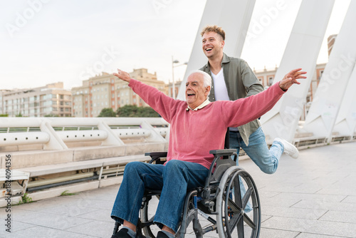 Laughing grandson pushing cheerful elderly man in wheelchair with arms raised on urban bridge