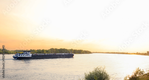Foto A long cargo vessel sailing on a wide river at golden hour, surrounded by calm water and lush green trees, highlighting peaceful inland waterway transport