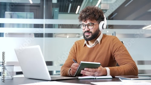 Businessman in headphones watching video call conference or training takes notes in a notebook looking at laptop screen sitting in business office. Worker listening to remote online course or seminar
