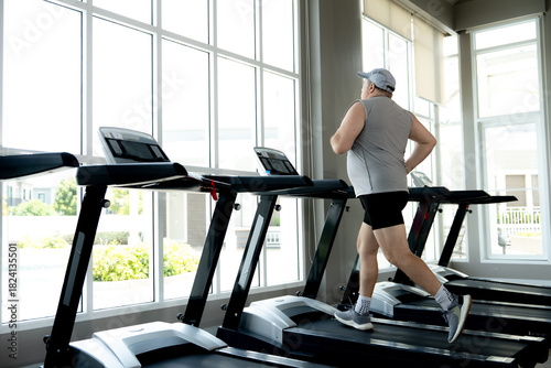 An active senior man is running on a treadmill at the fitness center. He enjoys his daily cardio workout indoors near a large window.
