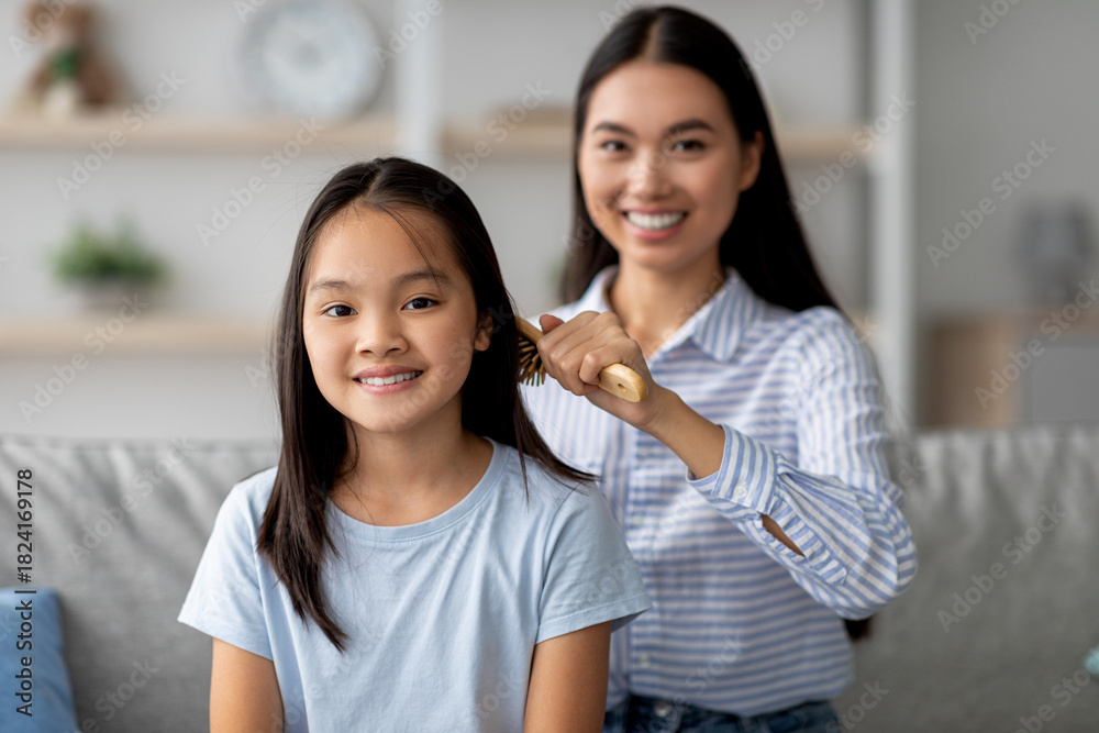 Fototapeta premium In a bright living room, a young mother gently combs her daughter's hair with a wooden brush. Both are relaxed on the sofa, sharing a tender moment filled with love and care.