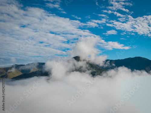 Aerial view of a sea of clouds partially obscuring the lush, undulating hills of the Phobjikha Valley under a bright blue sky, Nubding, Wangdue Phodrang, Bhutan.