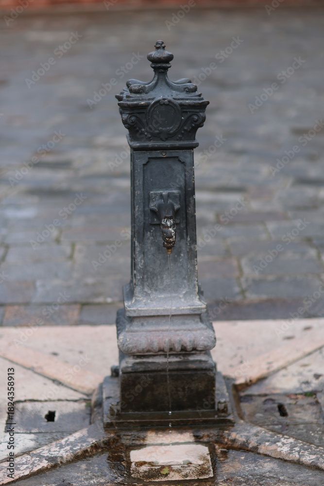 Obraz premium Old cast iron water fountain with decorative details and a small stream of water flowing, set on a stone pavement in Venice, Italy. Classic European street element.
