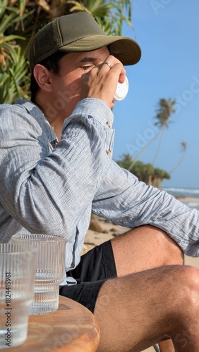 Serene peaceful relaxing escape for quiet contemplation and confident enjoyment as an unrecognizable man sits outside holding a coffee cup near the calm tropical ocean shoreline with palm, sand, sea