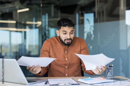 Confused businessman holding two documents, comparing data and reports while feeling stress and uncertainty about financial information and paperwork in office