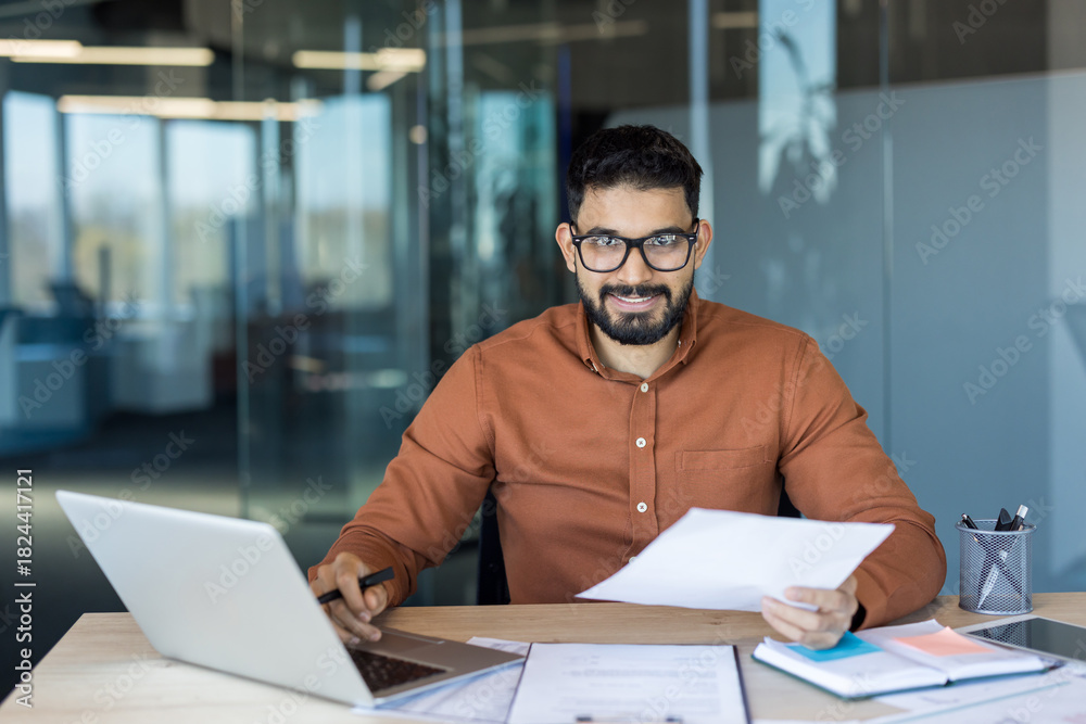 Fototapeta premium Indian businessman sitting at an office desk, smiling at the camera while holding documents and working on a laptop, managing tasks and paperwork in a modern workspace