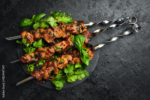 Delicious juicy skewers with pork meat grilling, grilled pork kebabs on a stone slate plate. Top view. On a dark textured background.