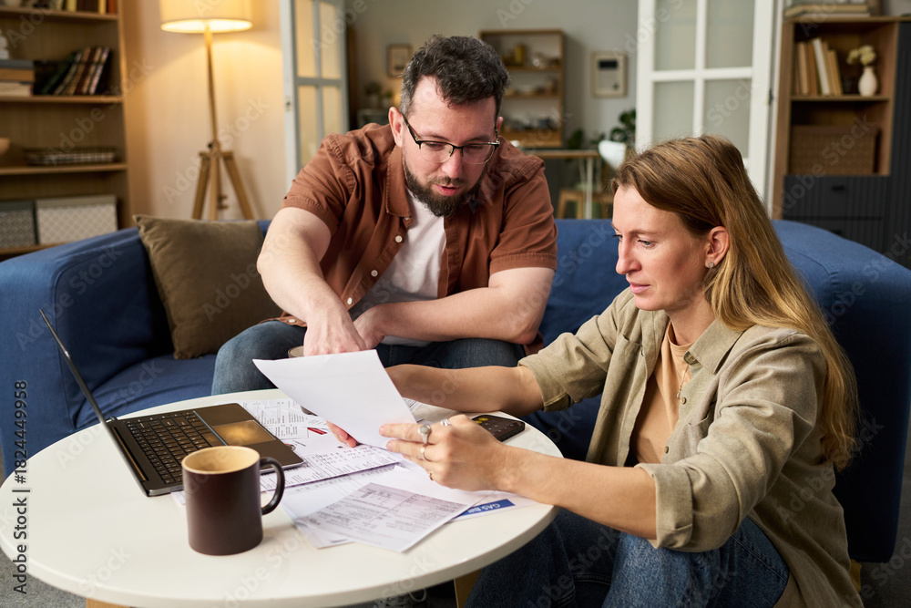 © pressmaster - Caucasian middle aged man and Caucasian middle aged woman reviewing tax documents together at table, man pointing at paper while woman holding paperwork, laptop and calculator visible
