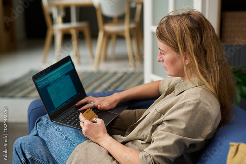 Caucasian middle aged woman sitting on sofa using laptop for online banking holding credit card preparing to pay taxes focusing on screen