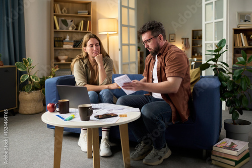 Middle aged Caucasian man explaining tax documents to middle aged Caucasian woman while sitting together at table with laptop and paperwork, both focused on financial planning