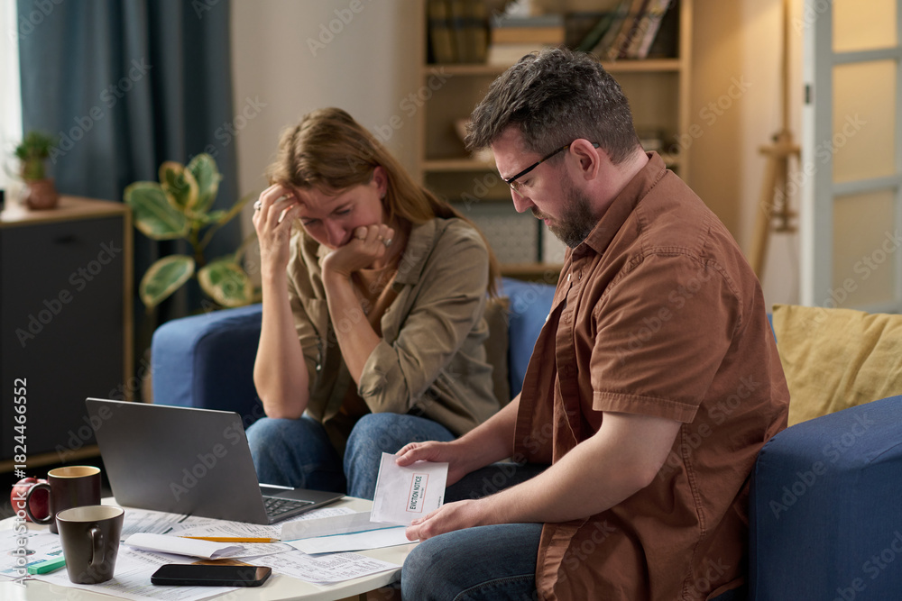 Obraz premium Middle aged Caucasian man holding tax document while sitting with worried middle aged Caucasian woman at table, both reviewing financial paperwork and using laptop for paying taxes