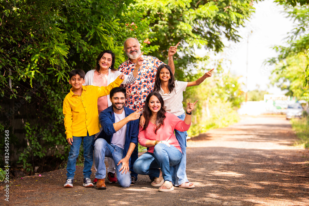 Fototapeta premium Happy Indian family of six sitting on lawn and pointing to present something outdoors.