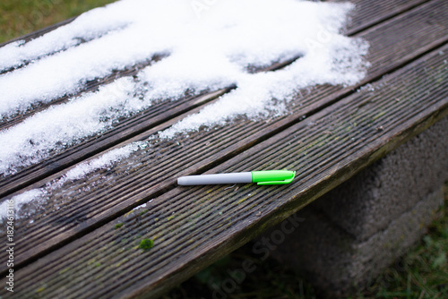 Fototapeta Naklejka Na Ścianę i Meble -  A green drawing marker lies on the corner of a street bench next to a small patch of snow in autumn.