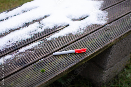 Fototapeta Naklejka Na Ścianę i Meble -  A red drawing marker lies on the corner of a street bench next to a small patch of snow in autumn.