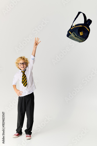 Excited schoolboy with wavy hair tosses backpack in a bright studio setting