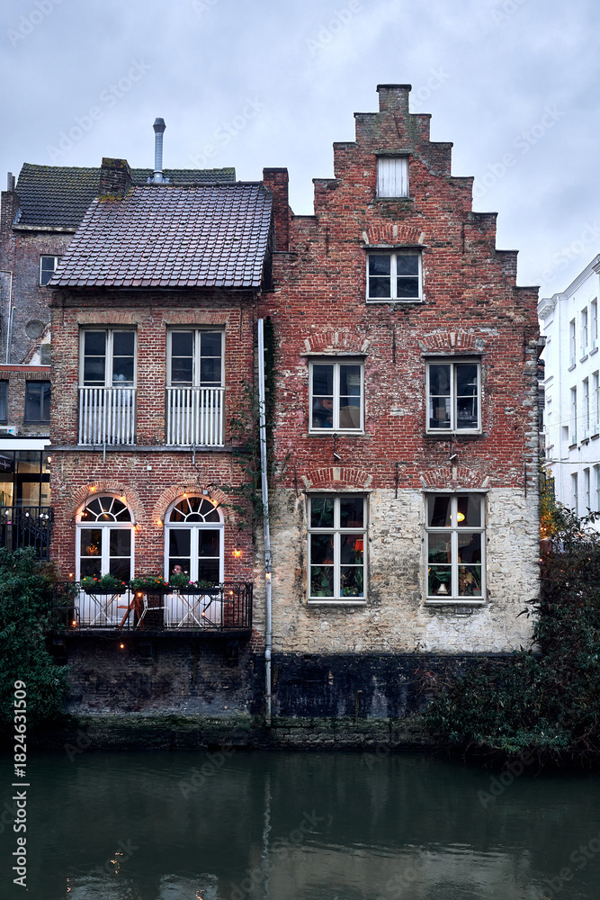 Fototapeta premium Two charming medieval buildings stand side by side on the banks of the River Lys in Ghent, Belgium. Their rustic red brick façades and simple windows overlook the canal