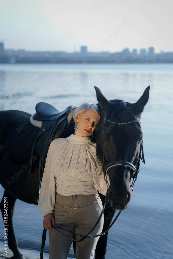 Obraz premium An adult woman holding a black horse and standing near the river