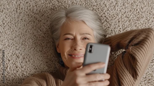 Smiling senior woman lies comfortably on soft carpet at home while adjusting her smartphone, shifting her gaze between the screen and her hand, maintaining a warm and relaxed expression