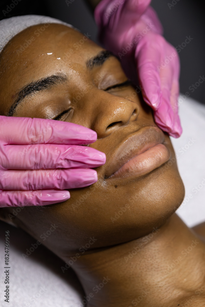 Naklejka premium African woman in beauty clinic doing a facial evaluation and a cosmetology treatment. Woman on an esthetician appointment of facial. use mask do skincare routine