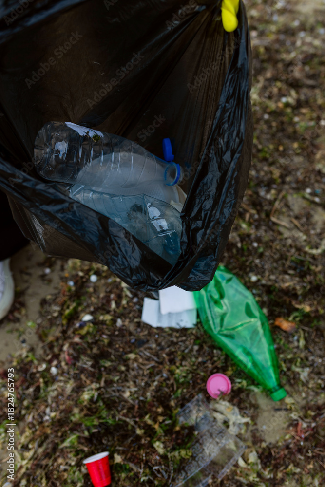 Obraz premium Volunteer collecting plastic waste and litter into a black trash bag during beach cleanup, highlighting pollution, environmental protection, sustainability, and eco awareness.