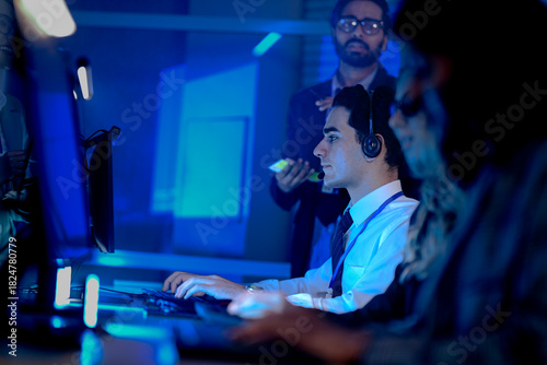 IT support team lead oversees a junior agent handling a complex technical issue. He offers real time advice while reviewing progress on screen in a busy operations center.