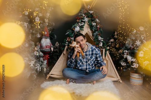 A smiling, dark-haired man with wrinkles on his face, wearing jeans and a shirt, sits near a Christmas tree, holding a stuffed animal. The man is surrounded by festive lights and a bokeh effect
