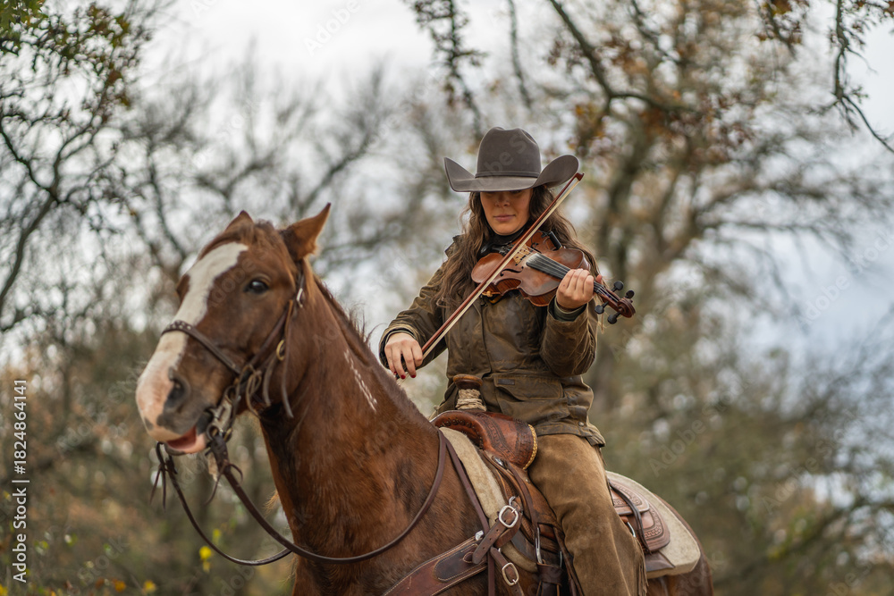 Fototapeta premium Cowgirl Woman Wearing Cowboy Hat Looking Down and Playing Fiddle Violin While Sitting On Brown and White Horse In Front of Trees and Cloudy Sky