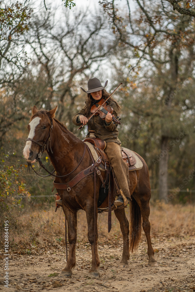 Fototapeta premium Cowgirl Woman Wearing Cowboy Hat Looking Down and Playing Fiddle Violin While Sitting On Brown and White Horse In Front of Trees and Cloudy Sky