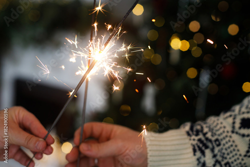 Hands holding fireworks against christmas tree and fireplace in festive decorated cozy living room. Happy New Year! Atmospheric family celebration with burning sparklers in hands close up