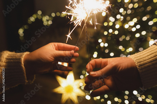 Hands holding fireworks against christmas tree lights bokeh in evening cozy living room. Happy New Year eve! Atmospheric family celebration with burning sparklers in hands close up