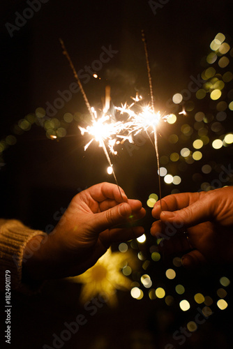 Hands holding fireworks against christmas tree lights bokeh in evening cozy living room. Happy New Year eve! Atmospheric family celebration with burning sparklers in hands close up