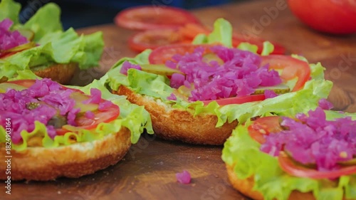 Several open buns with fresh lettuce, sliced tomatoes, and finely chopped pink onions on toasted bread. Hamburgers waiting for the patties to be cooked