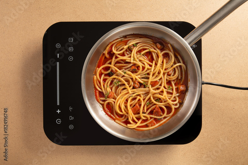 Long strands of spaghetti simmer in a rich tomato sauce in a stainless steel pot on an electric cooktop. The warm kitchen space invites a sense of comfort and home cooking.