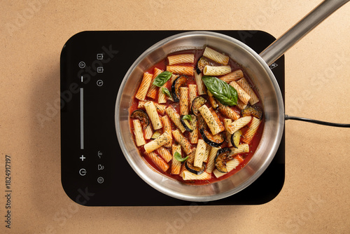 Pasta is being prepared in a stainless steel pot on a sleek induction stove. The dish features colorful vegetables and fresh basil, showcasing a vibrant meal in progress.
