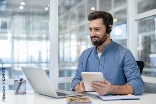 Happy bearded man wearing a headset and working remotely from an office, providing online customer support and technical assistance while looking at a laptop screen and holding a digital tablet