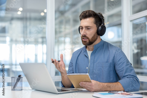 Man in headphones having a video conference call on a laptop, discussing and taking notes from an online meeting while working remotely from a modern office environment