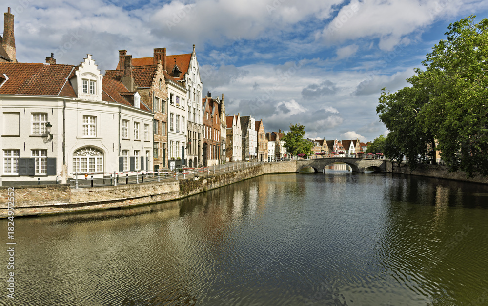 Obraz premium Row of traditional buildings along a canal in Bruges, Belgium, featuring stepped gables and facades in beige, white, and brick red. The calm canal reflects the architecture,