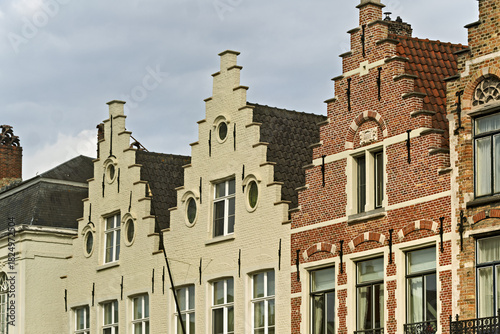 Group of historic buildings in Bruges, Belgium, showcasing steep red-tiled roofs, decorative brick chimneys, and gabled facades.