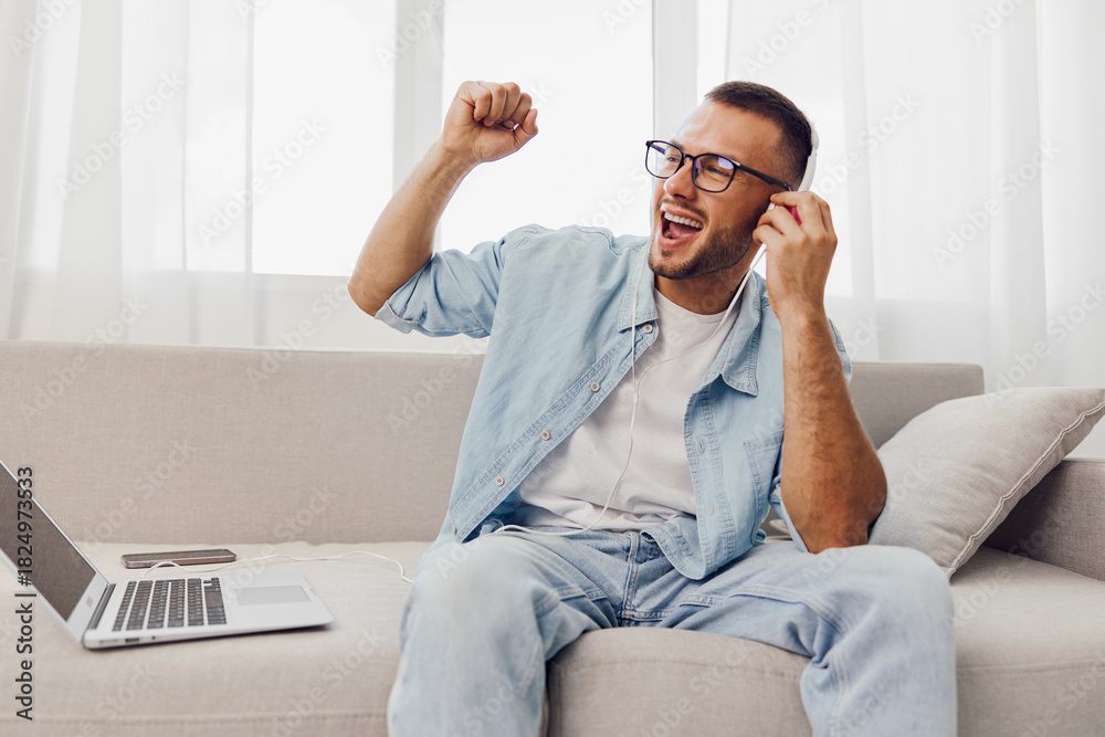 Fototapeta premium Energetic man enjoying music on headphones, sitting on a modern sofa with a laptop, celebrating his creative break. Perfect for showcasing relaxation and productivity.