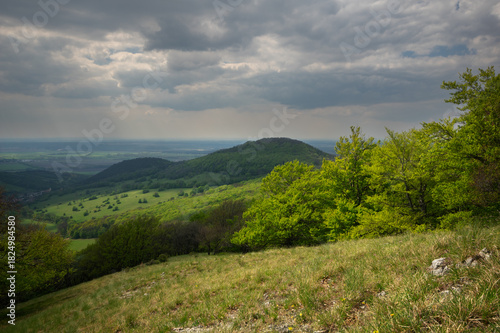 View of rolling green hills blanketed in vibrant spring foliage under a dramatic, cloud-strewn sky, Klokoc, Bratislava Region, Slovakia.
