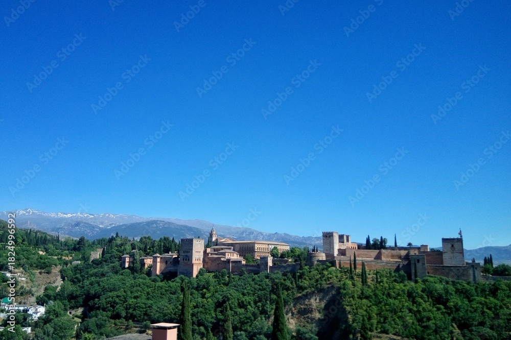 Obraz premium Alhambra Palace and Fortress complex on a forested hill with Sierra Nevada mountains and snow caps in the background under clear blue sky