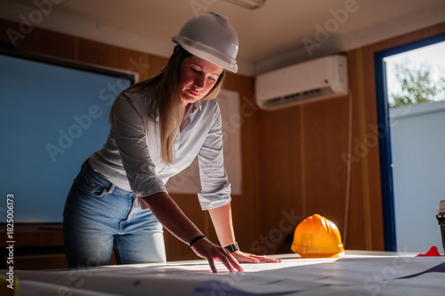 Young female architect wearing a white safety helmet, examining blueprints on a table in a temporary container office, focused on construction plans