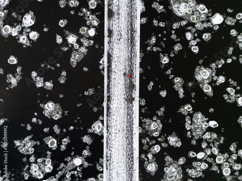 Aerial view of a solitary figure traversing a narrow bridge amidst the icy embrace of the Neris River's shuga, a dance of dark water and glimmering ice, Vilnius, Vilnius, Lithuania.