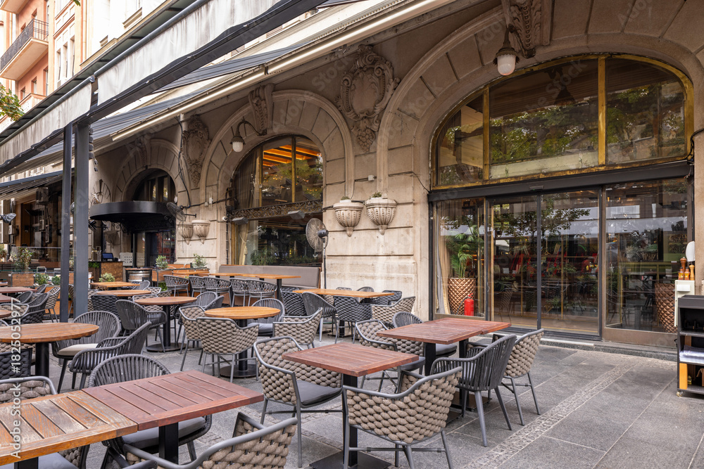 Naklejka premium Outdoor cafe terrace on a city street with tables, chairs, greenery, and large trees providing shade