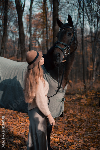 Foto Woman stands beside horse in forest during fall season while wearing a hat and s