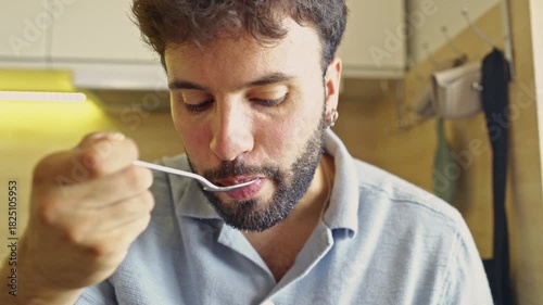A man with a beard sits in his modern kitchen, enjoying a casual breakfast with audible sounds. He takes a spoonful of food, chewing thoughtfully, as he starts his day.