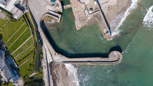 Aerial view of the historic Charlestown Harbour's sturdy stone quays embrace the tranquil, green-tinged waters against the crashing waves of the sea, Charlestown, Saint Austell, United Kingdom.
